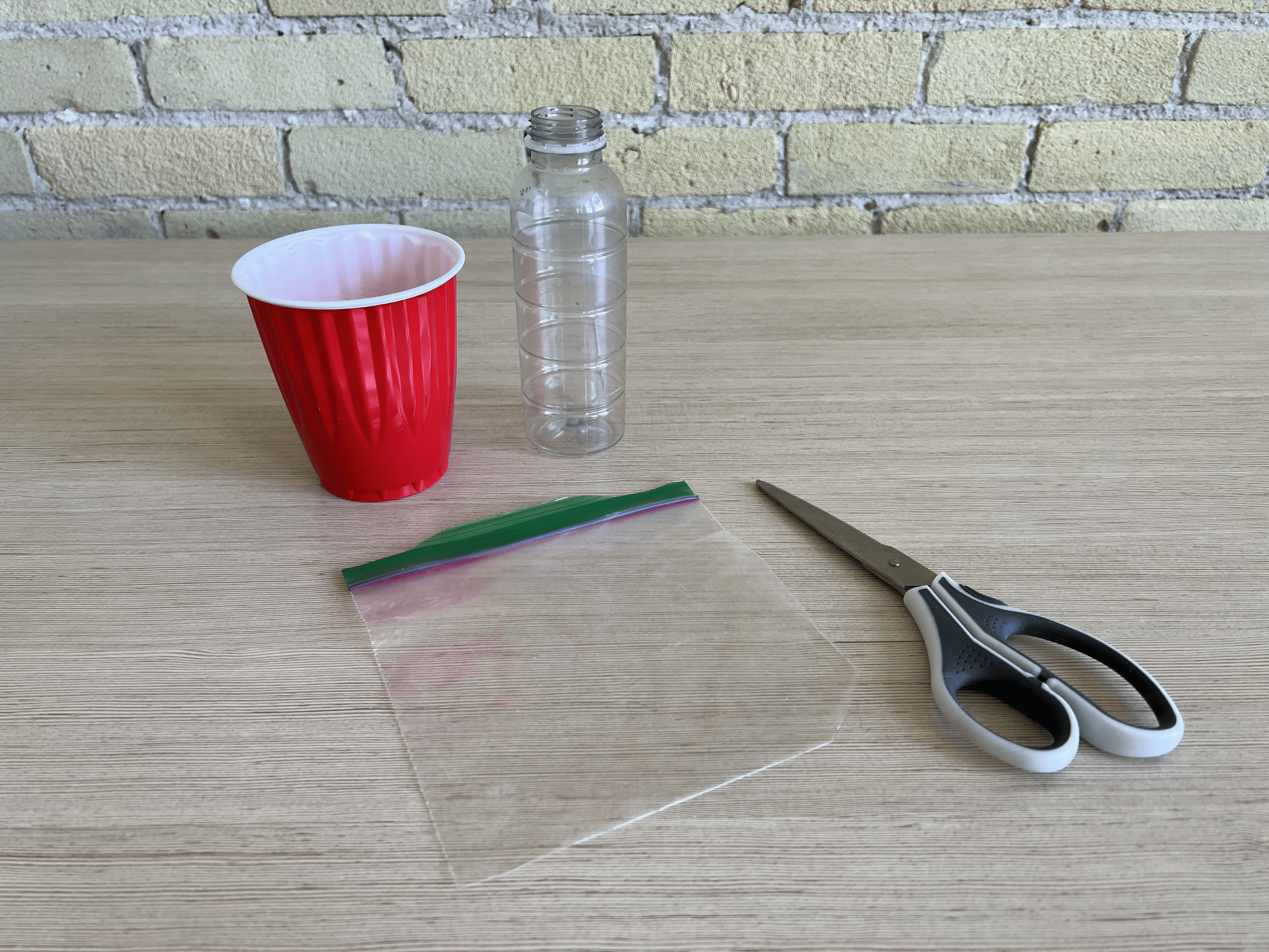 A ziploc bag with the corner cut off, a pair of scissors, a red solo cup, and an empty bottle sitting on a table.