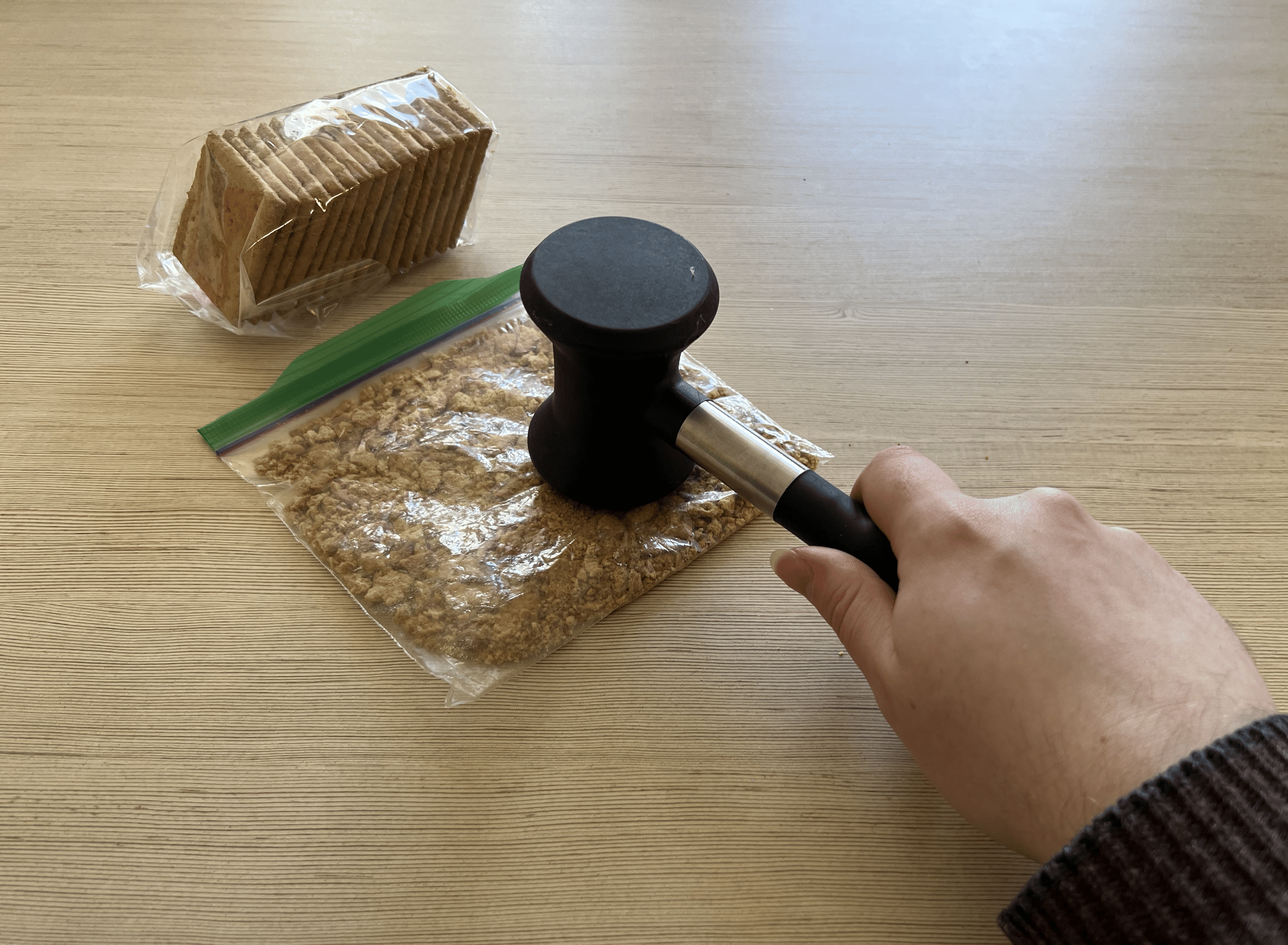 A hand holding the mallet and using it to crush up graham crackers in a ziploc bag.