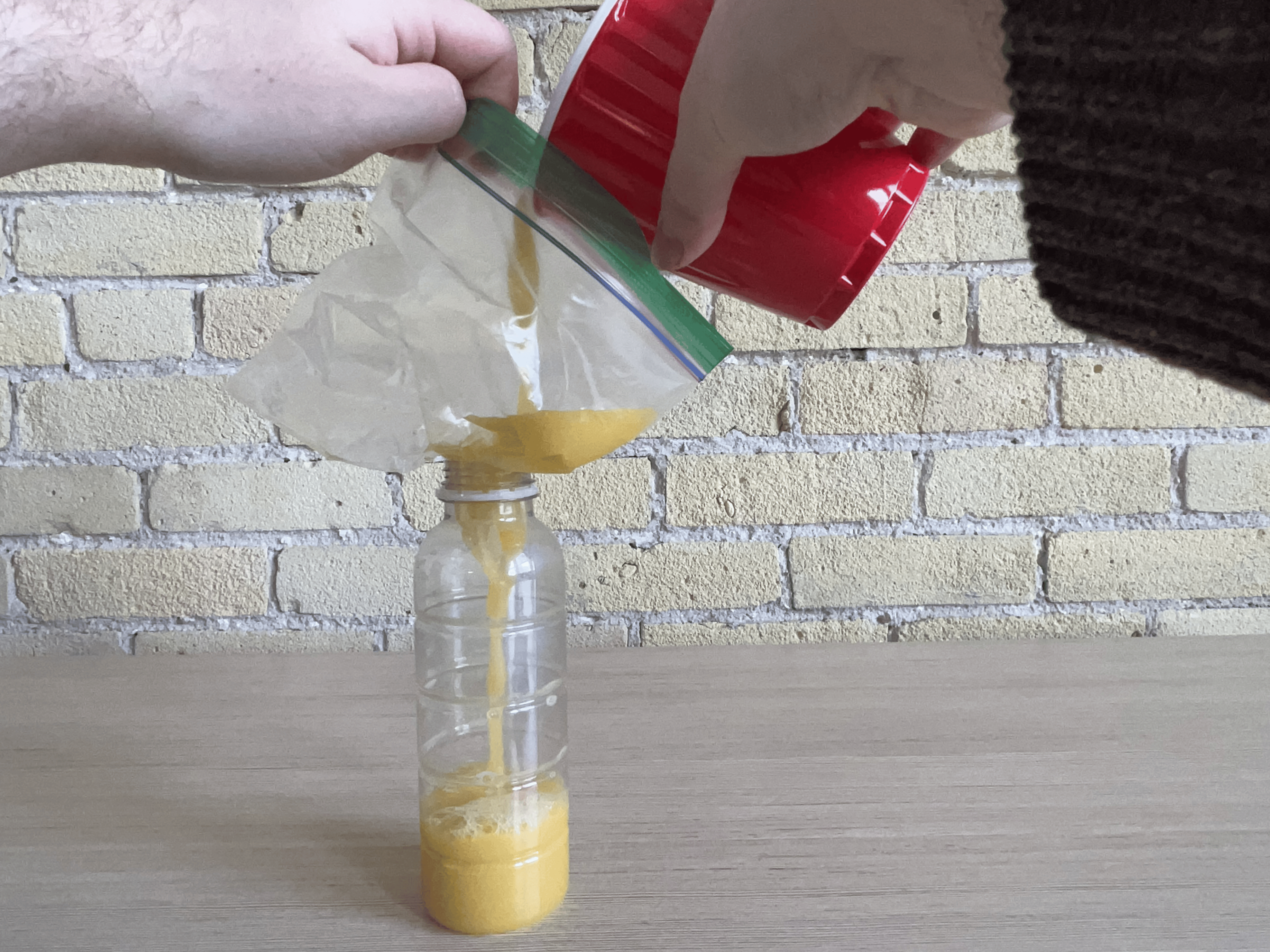 A pair o f hands pouring orange juice from the cup into the bottle using a ziploc bag as the funnel.