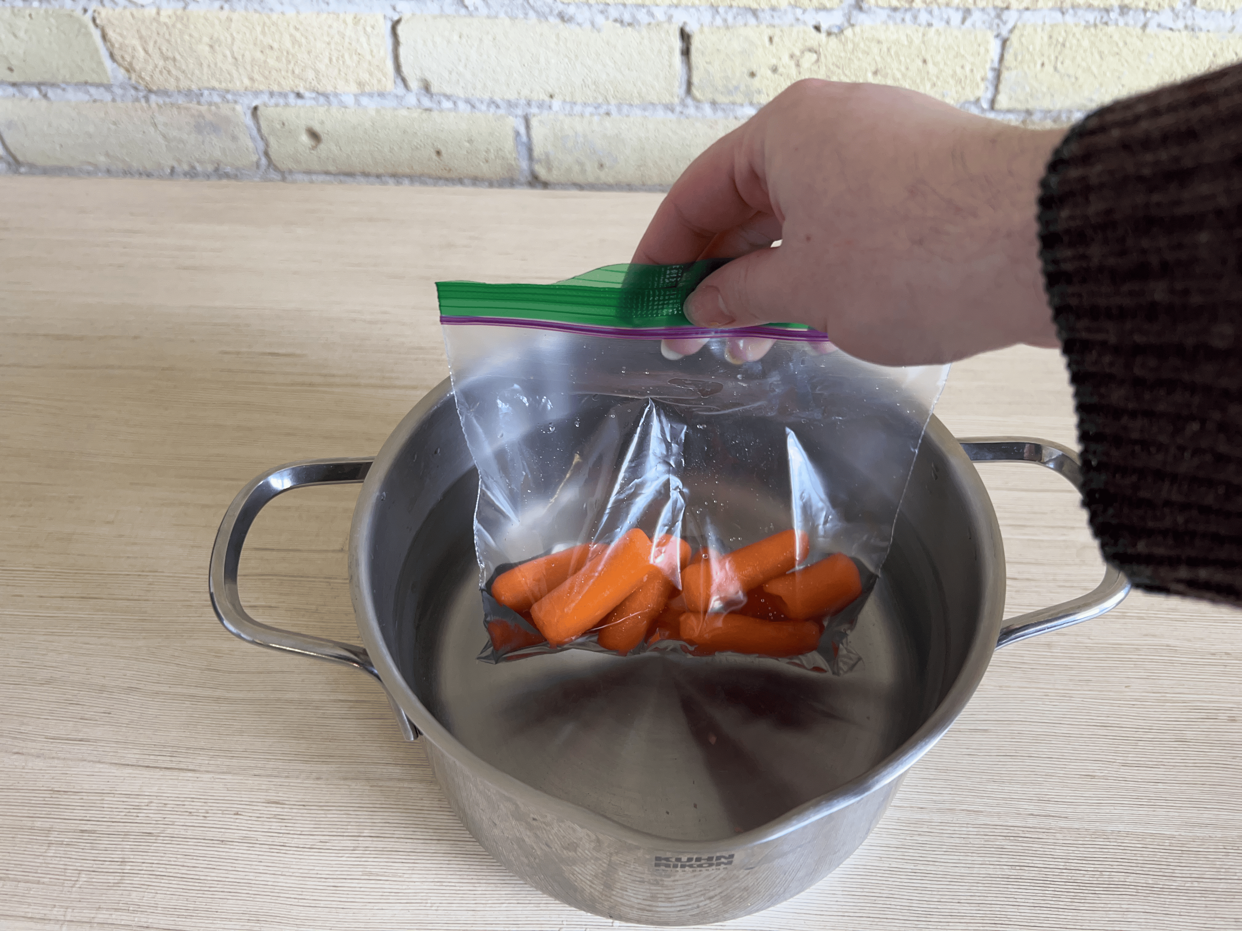 A hand dunking a ziploc bag of baby carrots into a pot of water.